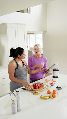 Dos-Mujeres-Mayores-Felices-Y-Diversas-Cortando-Frutas-Y-Usando-Tabletas-En-Una-Cocina-Soleada,-Cámara-Lenta