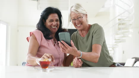 Felices-Y-Diversas-Mujeres-Mayores-Usando-Teléfonos-Inteligentes-Y-Discutiendo-En-Una-Cocina-Soleada,-Cámara-Lenta