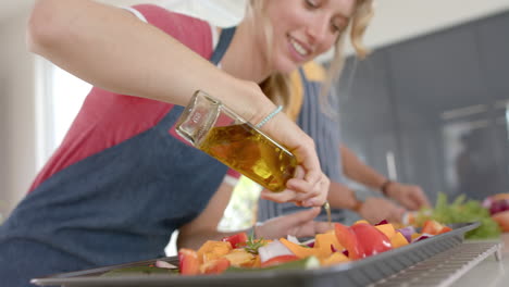 Happy-diverse-couple-preparing-fresh-vegetables-with-oil-and-wearing-aprons-in-kitchen,-slow-motion