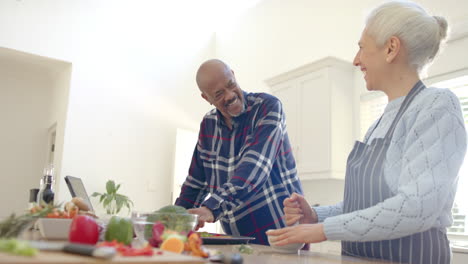 Happy-diverse-senior-couple-preparing-vegetables-and-using-tablet-in-kitchen,-slow-motion