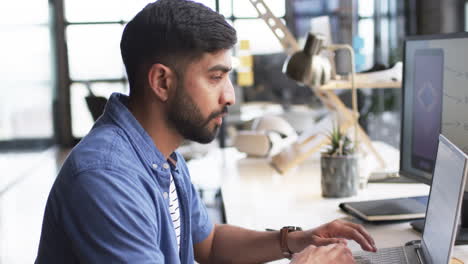 Young-Asian-man-with-beard-works-at-computer-in-office-setting,-indicating-business-environment