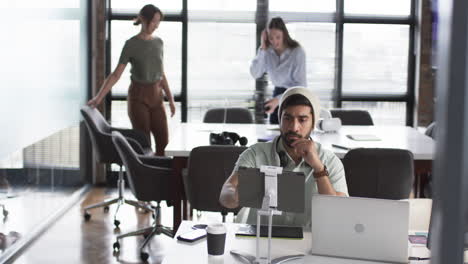 Young-Asian-man-in-casual-grey-beanie-sits-at-a-desk-with-a-laptop-in-a-business-office-setting