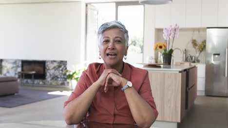 Biracial-woman-with-short-gray-hair-sits-at-a-table,-wearing-a-salmon-colored-dress