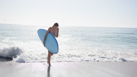 Junge-Kaukasische-Frau-Hält-Ein-Blaues-Surfbrett-An-Einem-Sonnigen-Strand-Mit-Kopierraum