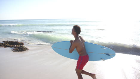 Un-Joven-Birracial-Lleva-Una-Tabla-De-Surf-Azul-Claro-En-Una-Playa-Soleada