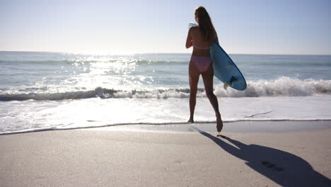 Una-Joven-Caucásica-Corre-Hacia-El-Océano-Con-Una-Tabla-De-Surf-Bajo-El-Brazo,-Con-Espacio-Para-Copiar