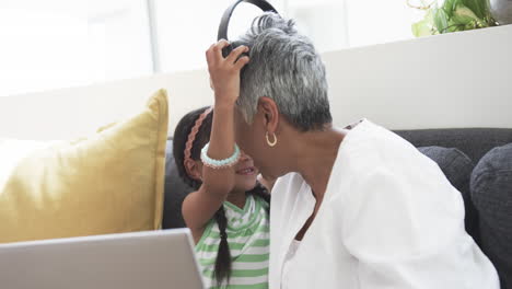 Biracial-girl-shares-headphones-with-her-grandmother,-both-smiling-in-a-home-setting