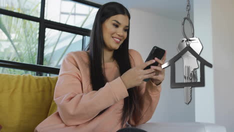 Animación-De-La-Llave-De-La-Casa-Plateada-Y-El-Llavero-Sobre-Una-Mujer-Birracial-Feliz-Usando-Un-Teléfono-Inteligente-En-Casa