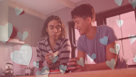 Animation-of-blue-hearts-over-happy-diverse-gay-male-couple-feeding-each-other-fruit-in-kitchen