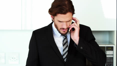 Businessman-talking-on-mobile-phone-while-using-laptop-in-kitchen