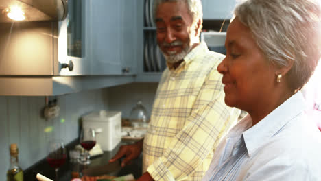 Un-Hombre-Mayor-Cortando-Verduras-Mientras-Una-Mujer-Usa-Una-Tableta-Digital-En-La-Cocina