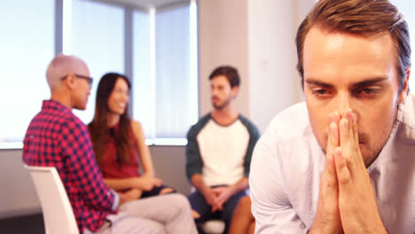 Upset-man-sitting-on-chair-with-colleagues-discussing-in-background