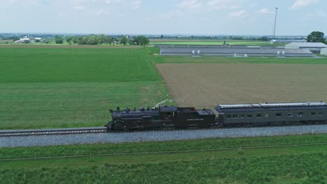 An-Side-Aerial-View-of-a-Steam-Passenger-Train,-Blowing-Smoke-and-Steam,-in-Slow-Motion,-on-a-Partially-Sunny-Day