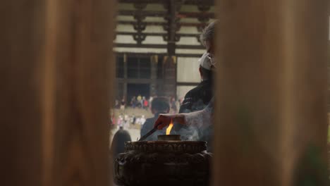 Hombre-Con-Mascarilla-Parado-Cerca-De-Un-Quemador-De-Incienso-Para-Encender-El-Palo-De-Incienso-Con-El-Gran-Salón-De-Buda-Del-Templo-Todaiji-Al-Fondo-En-Nara,-Japón