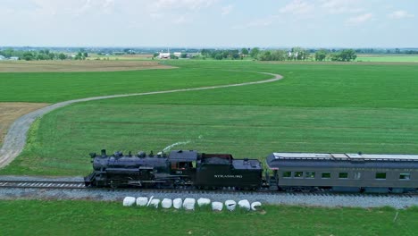 An-Side-Aerial-View-of-a-Steam-Passenger-Train,-Blowing-Smoke-and-Steam,-in-Slow-Motion,-on-a-Partially-Sunny-Day