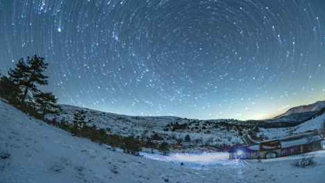 Star-trails-circling-in-the-night-sky-above-a-snowy-landscape-with-a-solitary-building-at-twilight,-winter-season,-long-exposure-timelapse