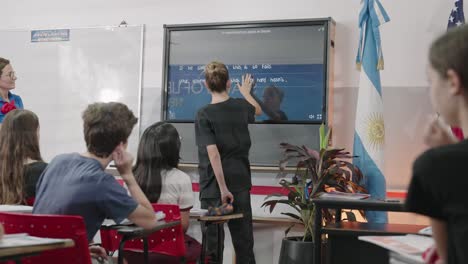 Moving-shot-of-a-teenage-student-in-a-classroom-with-other-students-doing-English-exercises-with-a-large-touch-screen