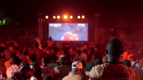 Hombre-De-Pie-Con-Camiseta-De-Costa-De-Marfil-Viendo-Un-Partido-Con-Aficionados-Al-Fondo,-Copa-Africana-De-Naciones-2023,-Abidján