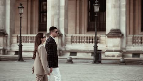 Stylish-couple-in-suits-wearing-sunglasses-walking-over-the-place-around-the-Museum-du-Louvre-in-Paris-France-with-its-baraque-buildings-without-tourists-around-showing-a-modern-classy-lifestyle