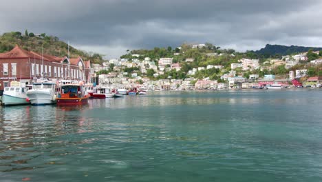 Colorful-boats-moored-at-Carenage-harbor-in-Grenada,-with-vibrant-houses-on-hillside,-cloudy-sky