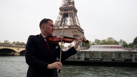 Ein-Kaukasischer-Geiger-In-Einem-Schwarzen-Anzug-Und-Mit-Roter-Krawatte-Spielt-Auf-Seiner-Geige-Vor-Dem-Berühmten-Eiffelturm-In-Paris,-Frankreich,-Während-Ein-Boot-Auf-Der-Seine-Vorbeischwimmt---Mittlerer-Rahmen