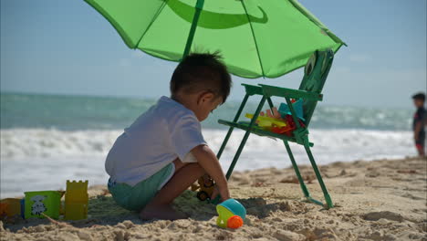 Zeitlupe-Eines-Süßen-Kleinen-Hispanischen-Jungen,-Der-An-Einem-Schönen,-Windigen-Und-Sonnigen-Tag-An-Der-Riviera-Maya-Mit-Seinem-Spielzeug-Neben-Seinem-Stuhl-Und-Sonnenschirm-Im-Sand-Spielt