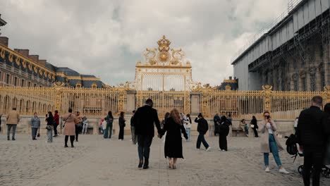Revealing-a-mixed-race-couple-walking-to-the-famous-Castle-Versailles-golden-main-gate-of-honour-with-tourists-around-on-a-cloudy-day---cinematic-wide-shot