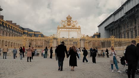 Revelando-Una-Pareja-De-Raza-Mixta-Caminando-Hacia-La-Famosa-Puerta-Principal-Dorada-Del-Castillo-De-Versalles-Con-Turistas-Alrededor-En-Un-Día-Nublado---Plano-Cinematográfico