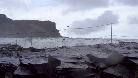 Rough-waves-crash-against-black-rocks-under-cloudy-skies,-cliff-in-the-background