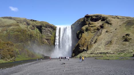 Touristen-Besuchen-Einen-Majestätischen-Wasserfall-In-Island-Mit-Einem-Sichtbaren-Regenbogen-An-Seiner-Basis,-Sonniger-Tag