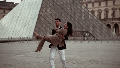 Elegant-couple-man-holding-his-women-on-hands-while-twirling-her-around-at-the-Museum-du-Louvre-Pyramid-surrounded-by-the-baroque-royal-residence-buildings-in-Paris-France---modern-lifestyle-and-suits