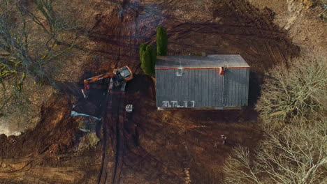 Aerial-drone-top-down-shot-over-crane-levelling-the-land-surrounding-a-wooden-house-along-rural-countryside-on-a-sunny-day