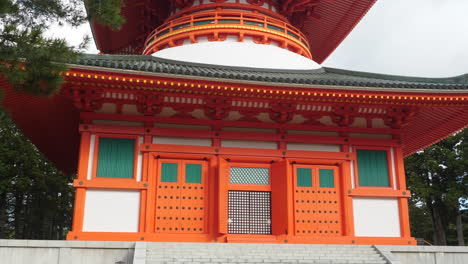 Gran-Pagoda-Naranja-Y-Blanca-En-Koyasan-Con-Intrincados-Detalles-Arquitectónicos,-Rodeada-De-Vegetación,-Bajo-Un-Cielo-Nublado,-Alejar