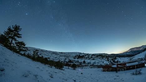 Twilight-over-a-snowy-landscape-with-stars,-a-rustic-barn,-and-trees,-capturing-the-serene-transition-from-night-to-day,-timelapse