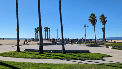 Schöner-Sommertag-Am-Venice-Beach-Mit-Leuten-In-Der-Ferne,-Die-Rollschuh-Laufen,-Vor-Blauem-Himmel-Und-Palmen