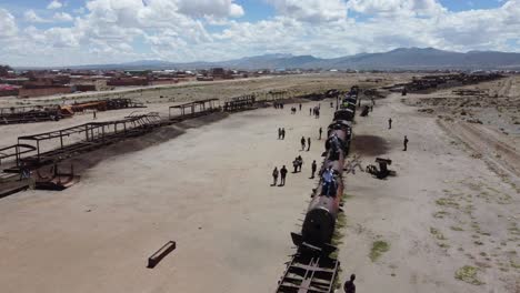 Tourists-visit-old-train-graveyard-in-Uyuni,-Bolivia,-aerial-flyover