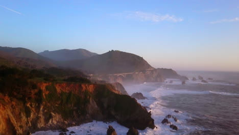 Bixby-Creek-Bridge-Big-Sur-California-aerial-cinematic-drone-flight-Pacific-Ocean-Nor-Cal-winter-summer-big-wave-swell-crashing-rugged-coastline-afternoon-golden-hour-pink-sunset-slowly-upward-motion