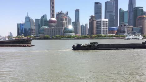 Möwe-Fliegt-Vor-Schiffen,-Die-Verschiedene-Güter-Und-Materialien-Auf-Dem-Huangpu-Fluss-In-Pudong,-Shanghai-Transportieren,-Wolkenkratzer-Im-Hintergrund
