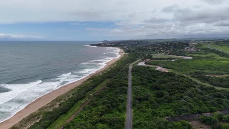 Aerial-of-the-beach-at-famous-divers-paradise-Aliwal-Shoal-Beach-near-Scottburgh,-South-Africa,-a-white-SUV-drives-down-a-rural-highway