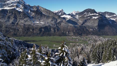 Fronalpstock-Glarus-Suiza-Vista-Aérea-Sobre-Un-Bosque-Nevado-Para-Revelar-Un-Pueblo-En-Un-Valle-Verde