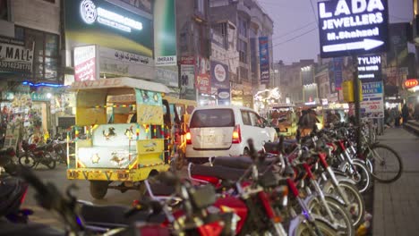 Busy-street-scene-in-Gujrat,-Pakistan-with-motorcycles,-rickshaw,-and-bustling-evening-crowd