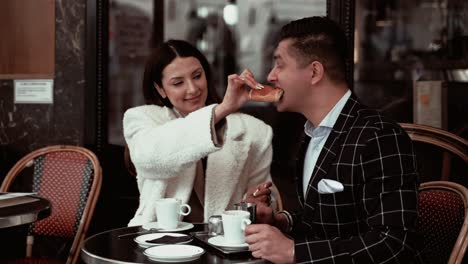 Elegante-Pareja-De-Cabello-Oscuro-Tomando-Un-Desayuno-Tradicional-Francés-Mientras-Las-Mujeres-Alimentan-A-Su-Marido-Con-Un-Croissant-De-Chocolate-Juguetón-Y-él-Está-Tomando-El-Bocado-En-Un-Café-Francés-En-París,-Francia