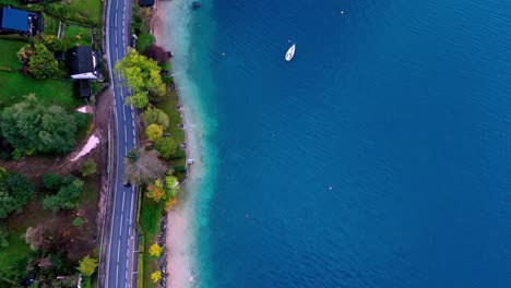 Luftaufnahme-Einer-Kurvenreichen-Straße-Entlang-Eines-Sees-Mit-Einem-Einsamen-Boot,-Das-Im-Stillen-Wasser-Vor-Anker-Liegt