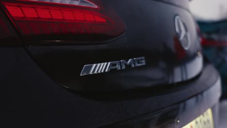 Close-up-of-black-Mercedes-Benz-car-rear-logo-and-AMG-letters,-people-walk-by