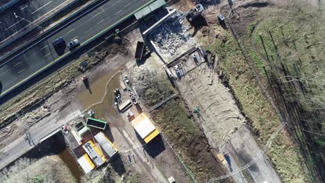 Drone-shot-of-a-construction-site-next-to-a-highway,-you-can-see-machinery-and-people-driving-on-the-highway