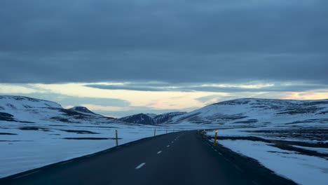 Driving-through-Iceland's-landscape,-snowy-mountains-in-view,-under-a-dawn-or-dusk-sky,-from-a-driver's-perspective