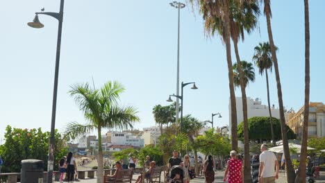 Tourists-and-locals-walking-by-the-bay-walk-in-Playa-De-Las-Americas-in-Tenerife,-dynamic-tilting-upwards-towards-palm-trees