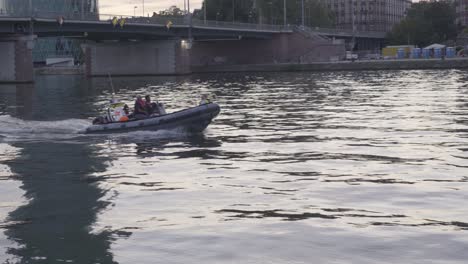 Inflatable-boat-with-crew-on-river-in-Frankfurt-at-dusk,-cityscape-and-bridge-in-background