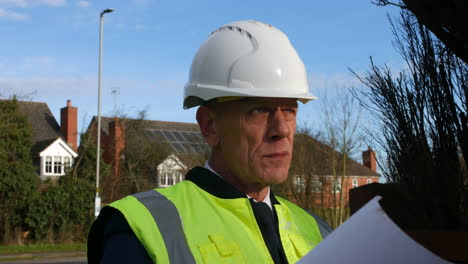 Close-up-of-a-senior-architect-electrician-tradesman-examining-plans-of-a-large-building-on-a-construction-site-in-a-residential-street-with-traffic-on-the-road-in-the-background