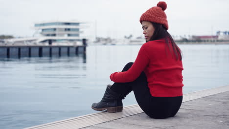 An-Asian-woman-relaxes-by-the-waters-of-Valencia-Port,-Spain,-wearing-a-red-beanie-that-complements-her-matching-checkered-jacket,-casting-a-serene-gaze-towards-the-camera